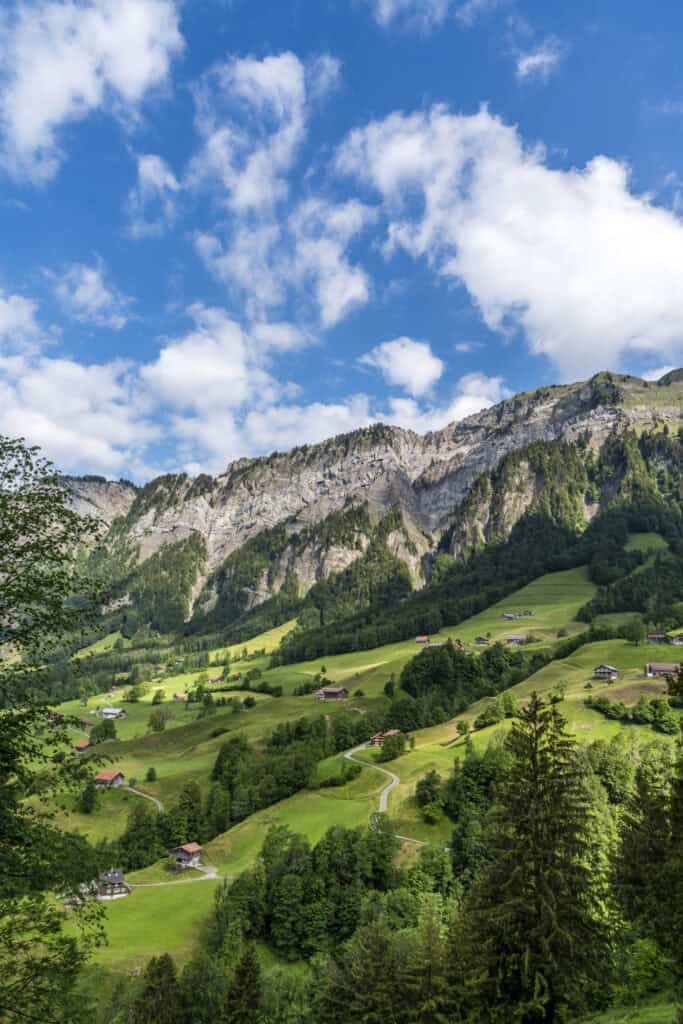 Berglandschaft mit grünen Hügeln, dramatischen Felsen, Ställen und Häusern und stahlblauer Himmel mit Quellwolken.