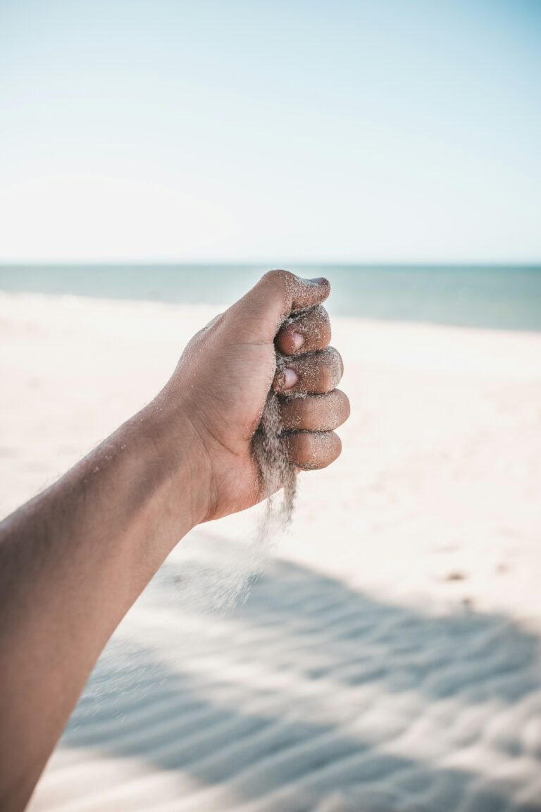 Hand vor Sandstrand mit Meer im Hintergrund, lässt Sand durch die Finger rinnen.