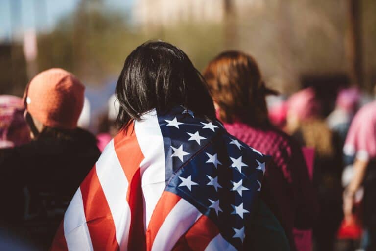 Menschen in einer Demonstration von hinten, eine Person hat eine amerikanische Flagge über den Schultern wie einen Umhang.